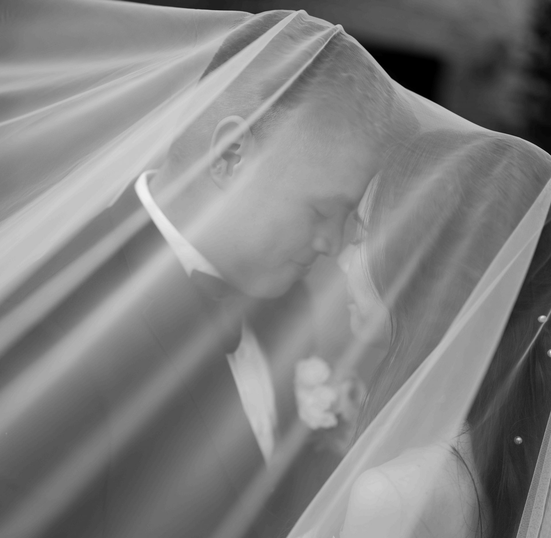 Gorgeous black and white photo of couple beneath veil, foreheads touching, eyes closed, and smiling.